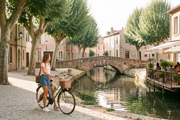 Une femme explore Castelsarrasin à pied avec un vélo, le long d’un canal bordé d’arbres, de bâtiments en briques et d’une terrasse de café.